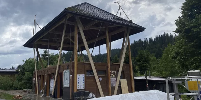Modernes Holzgebäude mit markantem, erhöhtem Satteldach und offener Konstruktion, aufgenommen an einem bewölkten Tag inmitten einer grünen Hügellandschaft.