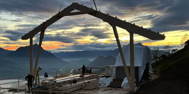 Holzbauarbeiten auf einer Bergbaustelle bei Sonnenaufgang – Zimmerleute montieren eine Dachkonstruktion mit Blick auf die umliegenden Berge und das Tal unter einem farbenprächtigen Morgenhimmel.