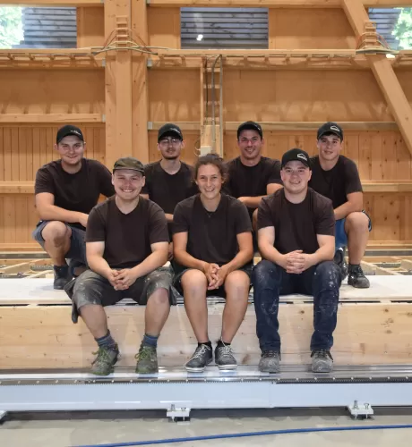Sieben junge Zimmerleute in Arbeitskleidung posieren lächelnd in einer Werkhalle. Drei Personen sitzen auf einer Holzkonstruktion, vier knien oder stehen dahinter. Alle tragen dunkle T-Shirts und teils Kappen, im Hintergrund ist eine moderne Holzbauhalle sichtbar.