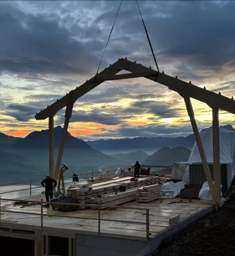 Holzbauarbeiten auf einer Bergbaustelle bei Sonnenaufgang – Zimmerleute montieren eine Dachkonstruktion mit Blick auf die umliegenden Berge und das Tal unter einem farbenprächtigen Morgenhimmel.