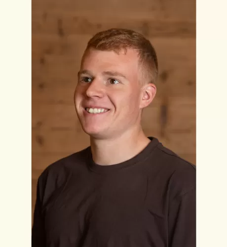 Porträt eines jungen Mannes mit kurzem, hellbraunem Haar, der lächelnd leicht nach rechts blickt. Er trägt ein dunkles T-Shirt, im Hintergrund ist eine warme Holzverkleidung zu sehen.