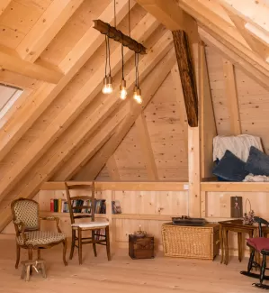 Gemütlicher Dachraum mit sichtbarer Holzbalkendecke, Holzboden und Einrichtung aus Stühlen, Bücherregal und warmem Licht.