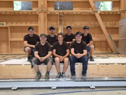Sieben junge Zimmerleute in Arbeitskleidung posieren lächelnd in einer Werkhalle. Drei Personen sitzen auf einer Holzkonstruktion, vier knien oder stehen dahinter. Alle tragen dunkle T-Shirts und teils Kappen, im Hintergrund ist eine moderne Holzbauhalle sichtbar.