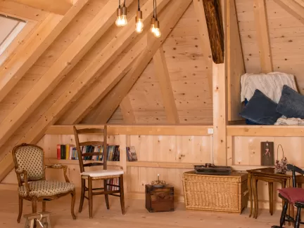 Gemütlicher Dachraum mit sichtbarer Holzbalkendecke, Holzboden und Einrichtung aus Stühlen, Bücherregal und warmem Licht.