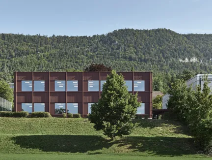 Aussenansicht eines modernen, zweigeschossigen Gebäudes mit dunkelbrauner Fassade und grossen, regelmässig angeordneten Fenstern. Das Gebäude steht in einer grünen Umgebung mit Wiese und Bäumen. Im Hintergrund erhebt sich ein bewaldeter Hügel unter klarem Himmel, was die ruhige, naturnahe Lage des Baus betont.