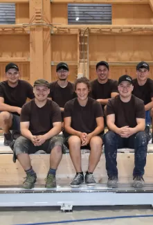 Sieben junge Zimmerleute in Arbeitskleidung posieren lächelnd in einer Werkhalle. Drei Personen sitzen auf einer Holzkonstruktion, vier knien oder stehen dahinter. Alle tragen dunkle T-Shirts und teils Kappen, im Hintergrund ist eine moderne Holzbauhalle sichtbar.