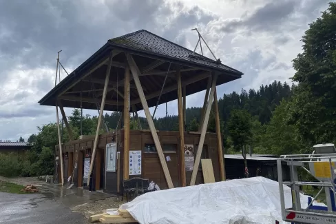 Modernes Holzgebäude mit markantem, erhöhtem Satteldach und offener Konstruktion, aufgenommen an einem bewölkten Tag inmitten einer grünen Hügellandschaft.