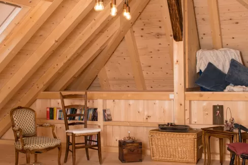 Gemütlicher Dachraum mit sichtbarer Holzbalkendecke, Holzboden und Einrichtung aus Stühlen, Bücherregal und warmem Licht.