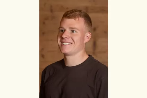 Porträt eines jungen Mannes mit kurzem, hellbraunem Haar, der lächelnd leicht nach rechts blickt. Er trägt ein dunkles T-Shirt, im Hintergrund ist eine warme Holzverkleidung zu sehen.