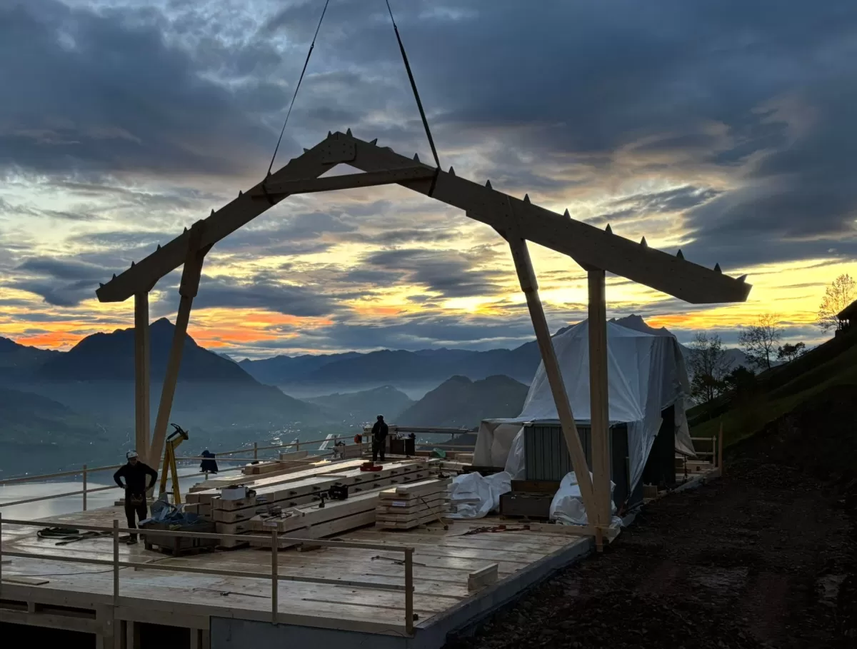 Holzbauarbeiten auf einer Bergbaustelle bei Sonnenaufgang – Zimmerleute montieren eine Dachkonstruktion mit Blick auf die umliegenden Berge und das Tal unter einem farbenprächtigen Morgenhimmel.