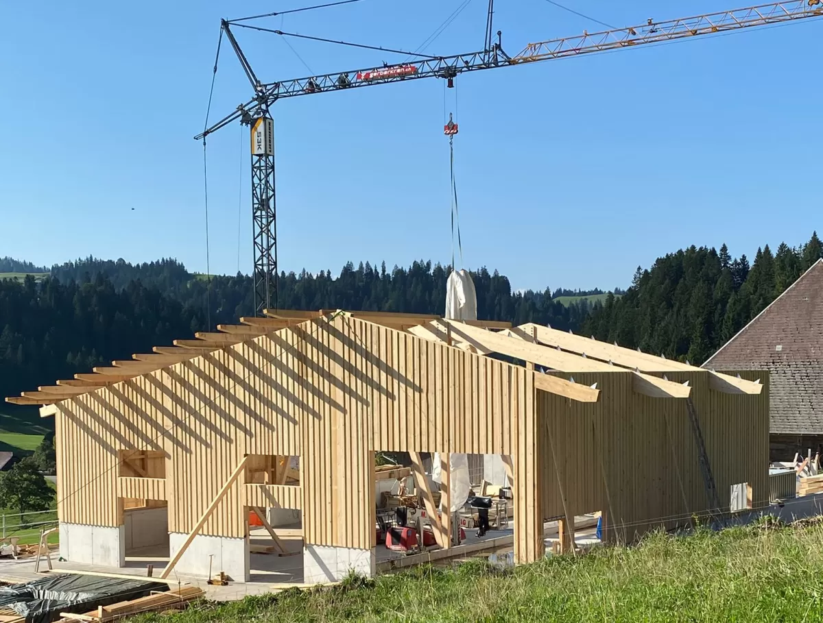 Ein modernes Holzbauprojekt von Zimmerei Hirschi AG: Im Vordergrund ist ein neu errichtetes Gebäude mit tragender Holzkonstruktion zu sehen. Ein Baukran hebt Materialien auf das Dach, während die klare Struktur der Holzbalken und Fassaden die Handwerkskunst hervorhebt. Umgeben von grünen Wiesen und bewaldeten Hügeln steht der Bau für nachhaltige, präzise und regionale Zimmermannsarbeit.