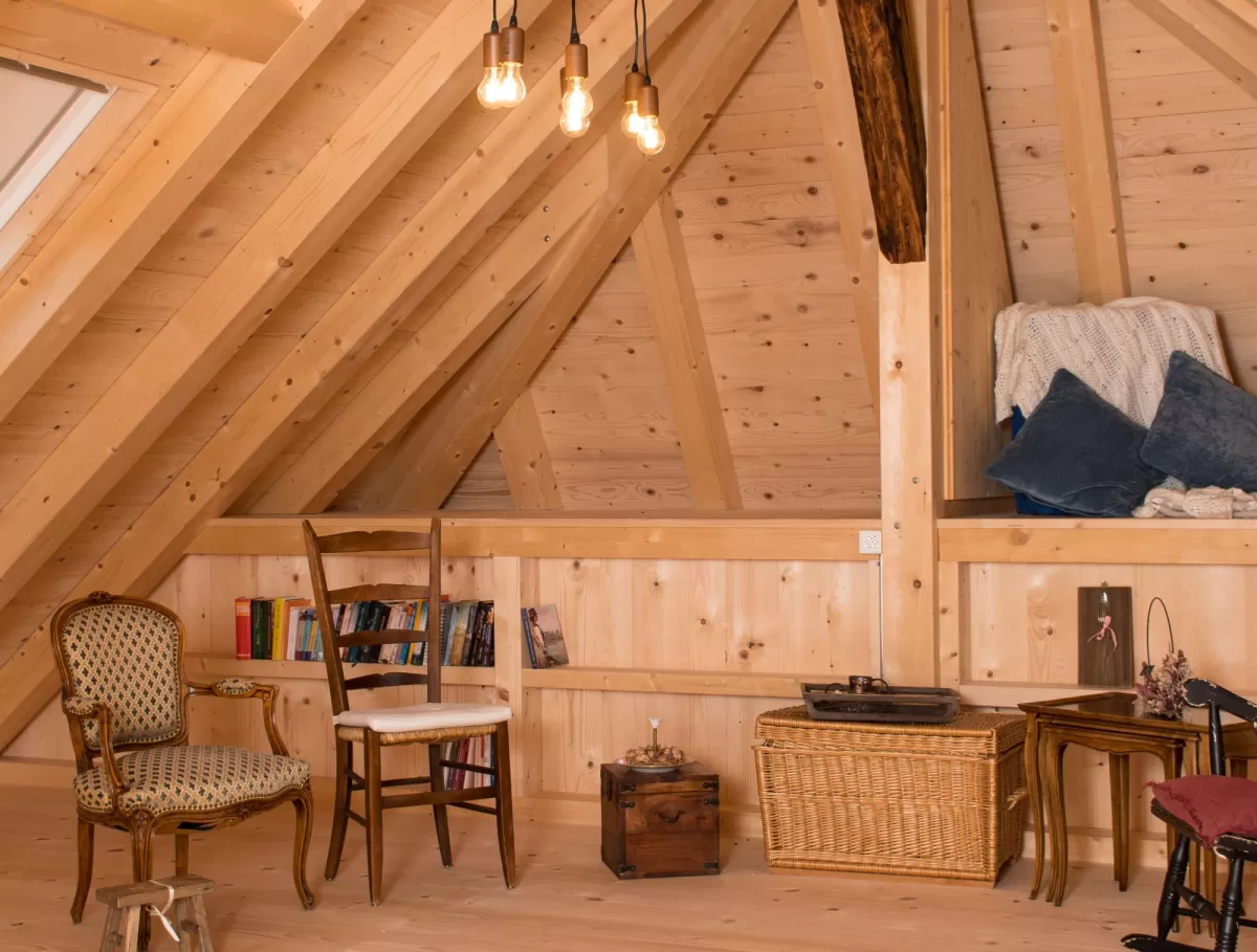 Gemütlicher Dachraum mit sichtbarer Holzbalkendecke, Holzboden und Einrichtung aus Stühlen, Bücherregal und warmem Licht.