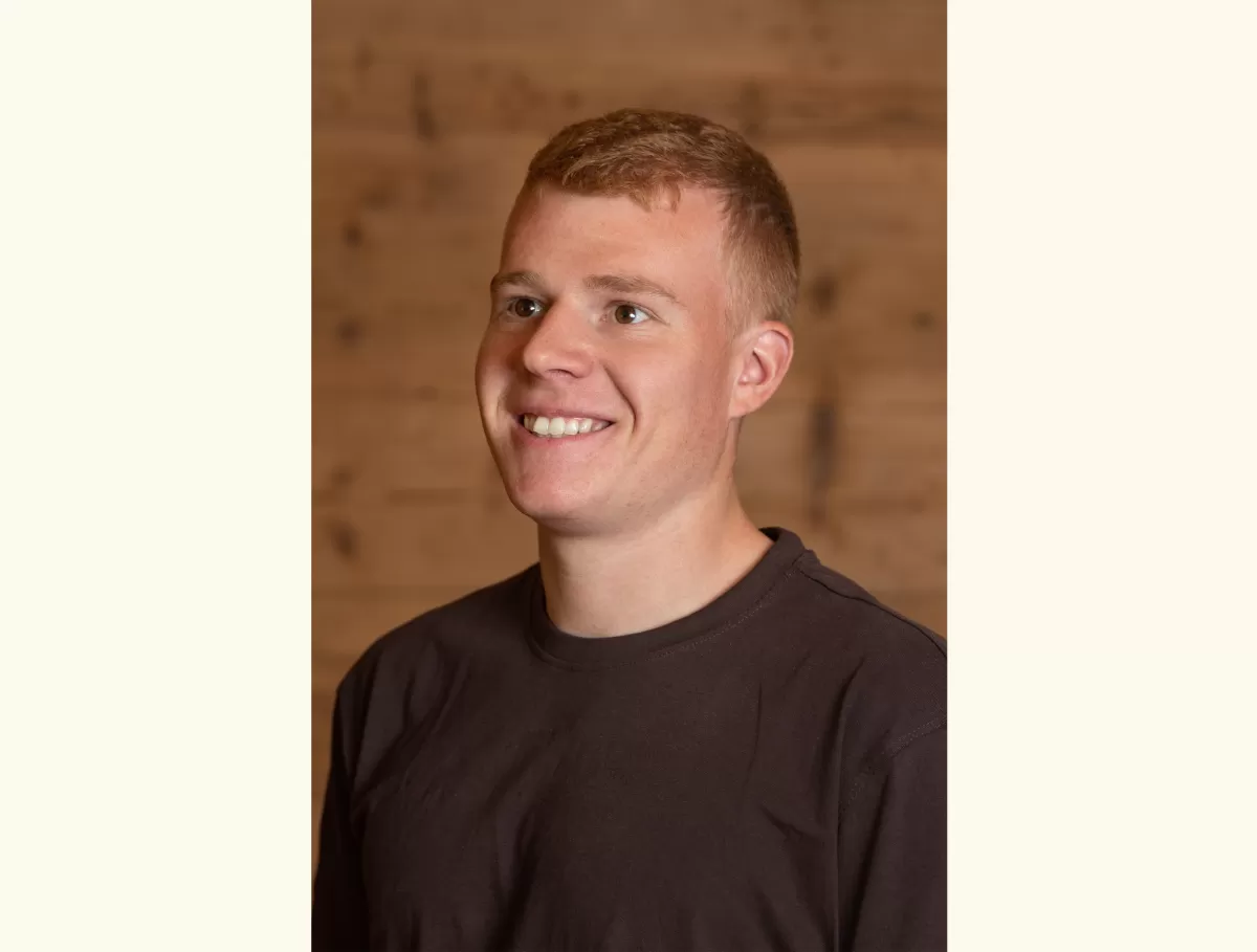 Porträt eines jungen Mannes mit kurzem, hellbraunem Haar, der lächelnd leicht nach rechts blickt. Er trägt ein dunkles T-Shirt, im Hintergrund ist eine warme Holzverkleidung zu sehen.