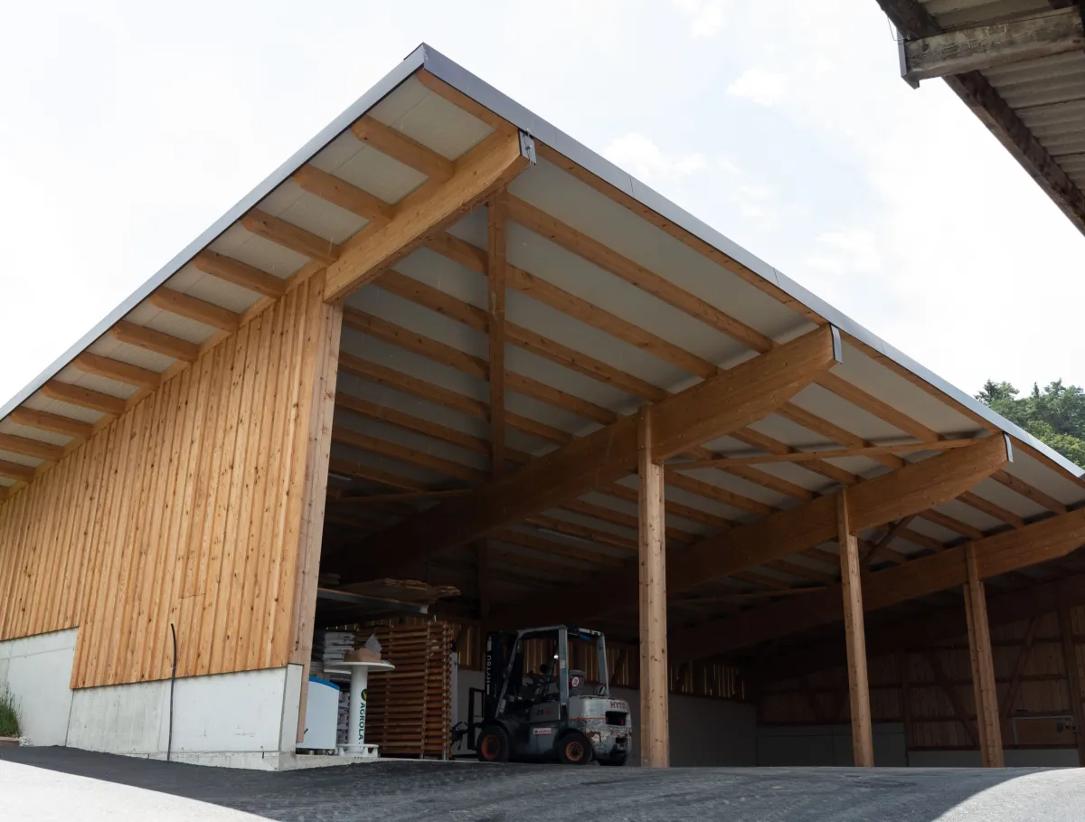 Aussenansicht einer grossen, offenen Lager- oder Maschinenhalle in Holzbauweise mit geneigtem Dach. Die Konstruktion aus sichtbaren Holzbalken ruht auf einem Betonsockel. Im Inneren der Halle sind ein Gabelstapler und gelagerte Materialien zu erkennen. Die Umgebung wirkt ländlich und funktional gestaltet.