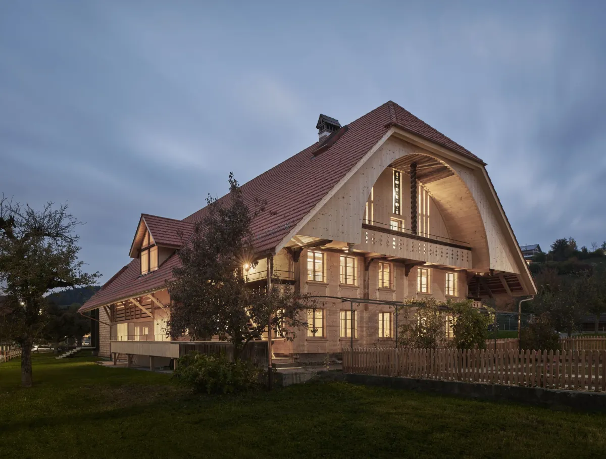 Traditionelles Holzhaus im Emmental in der Abenddämmerung, mit geschwungenem Giebeldach und aufwendig gestalteter Holzfassade. Warmes Licht fällt aus den Fenstern und betont die handwerklichen Details der historischen Holzkonstruktion. Das Gebäude steht in einer ruhigen, ländlichen Umgebung mit Garten, Bäumen und Holzzaun.