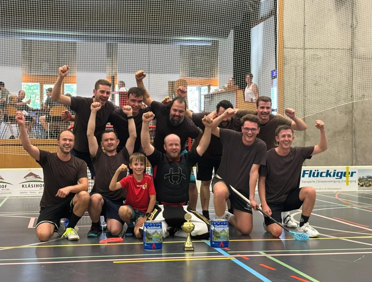 Eine Unihockey-Mannschaft feiert gemeinsam mit einem Kind und einem Torhüter auf dem Hallenboden einen Turniersieg. Vor der Gruppe stehen ein Pokal und zwei Getränkepreise.