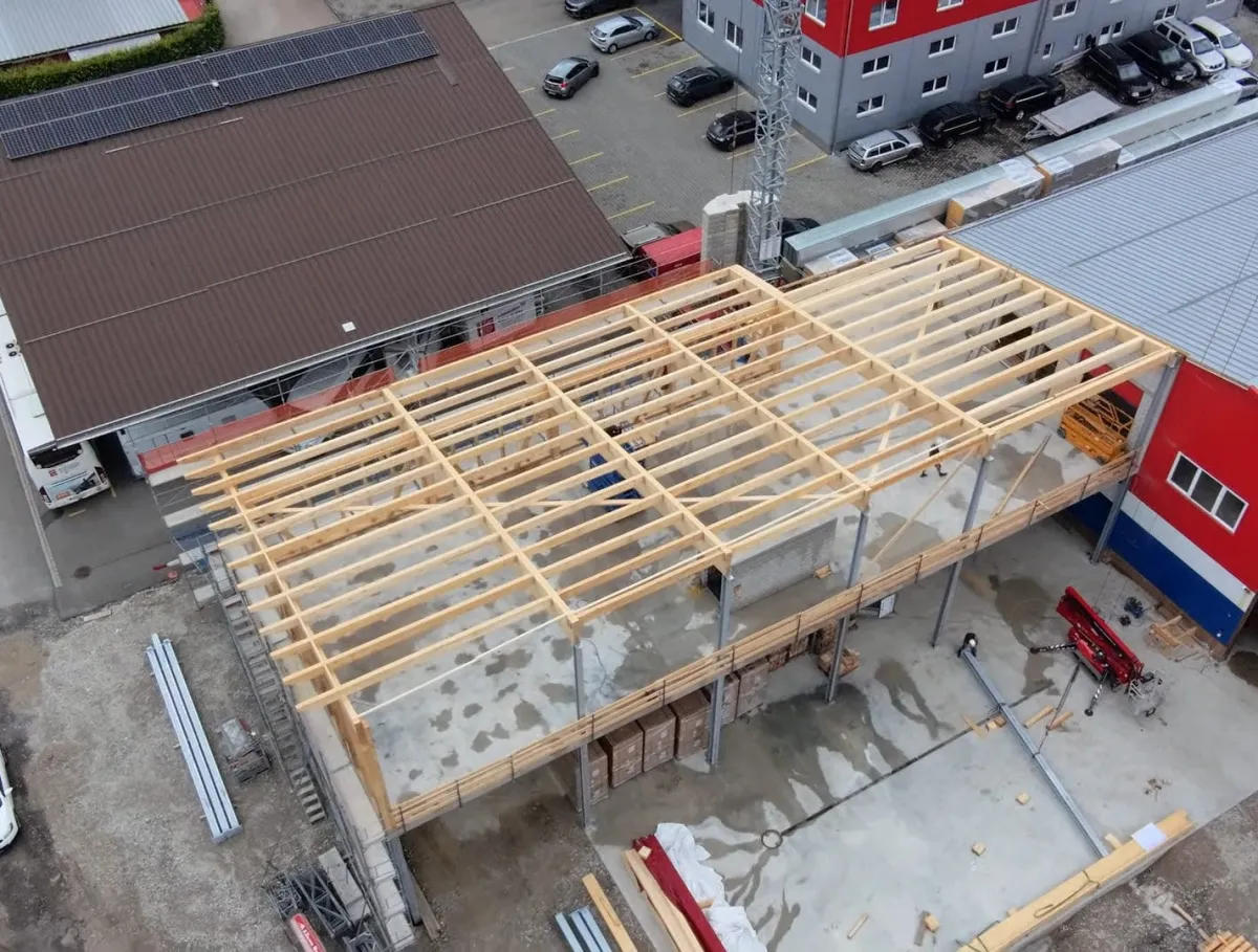 Luftaufnahme eines Gewerbebaus während der Bauphase. Zu sehen ist die im Aufbau befindliche Holzdachkonstruktion mit Tragwerk und Sparren auf einem Industriegebäude, umgeben von Werkhallen und Baustelleneinrichtungen.