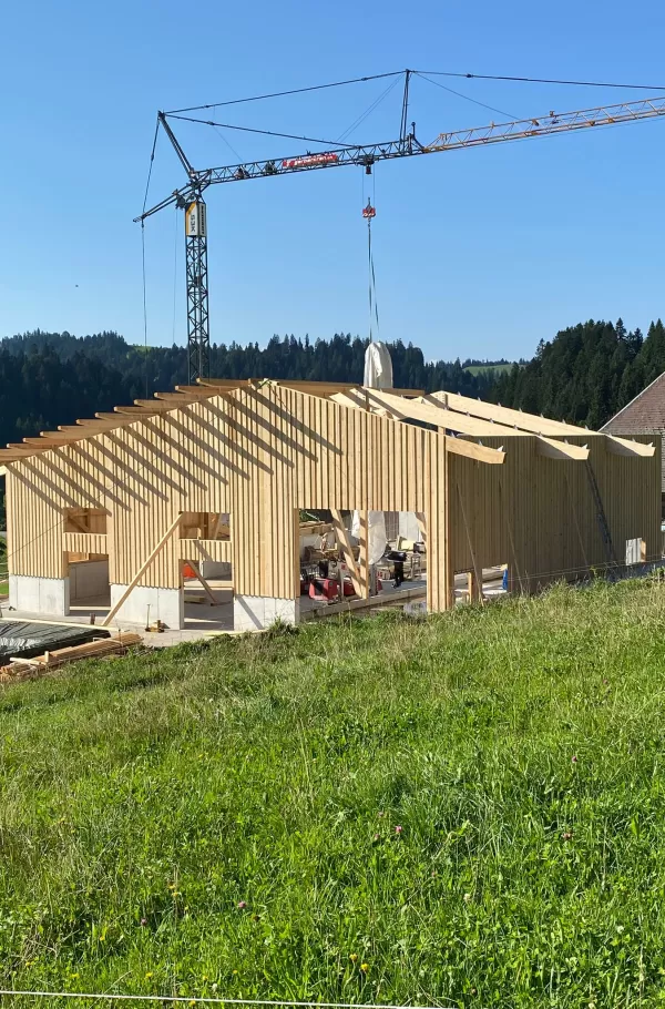 Ein modernes Holzbauprojekt von Zimmerei Hirschi AG: Im Vordergrund ist ein neu errichtetes Gebäude mit tragender Holzkonstruktion zu sehen. Ein Baukran hebt Materialien auf das Dach, während die klare Struktur der Holzbalken und Fassaden die Handwerkskunst hervorhebt. Umgeben von grünen Wiesen und bewaldeten Hügeln steht der Bau für nachhaltige, präzise und regionale Zimmermannsarbeit.