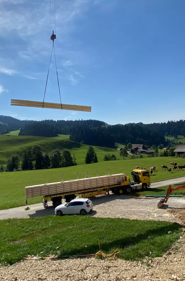 Ein Kran hebt frische Holzbalken hoch über die Baustelle, während im Hintergrund ein Lastwagen mit weiteren Holzelementen bereitsteht. Die Szene spielt inmitten der idyllischen Hügellandschaft des Emmentals, umgeben von grünen Wiesen und weidenden Kühen – ein Zusammenspiel von traditioneller Natur und moderner Holzbaukunst.