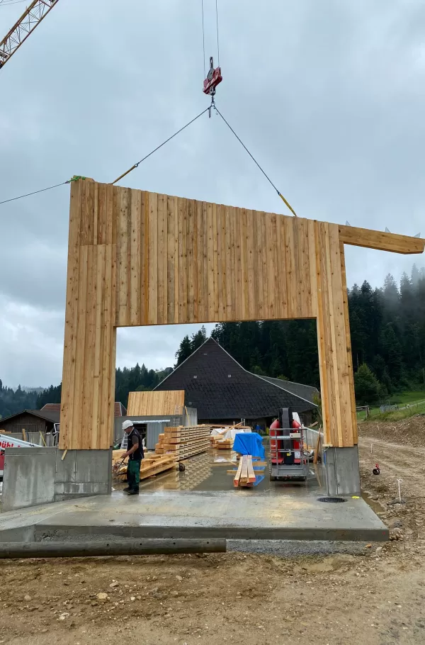 Eine massive Holzwand wird mit Hilfe eines Baukrans millimetergenau aufgestellt. Auf dem stabilen Betonfundament entsteht Schritt für Schritt ein neues Holzgebäude. Ein Zimmermann überwacht den Baufortschritt, während rundherum Bauholz und Geräte bereitliegen. Die Szene verdeutlicht Präzision, Teamarbeit und das Zusammenspiel von traditionellem Handwerk und moderner Technik.