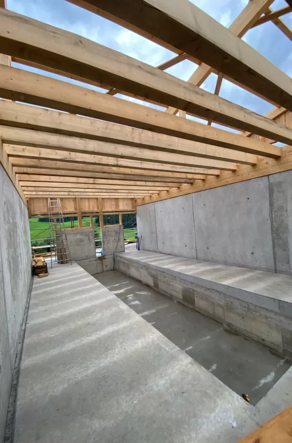Innenansicht eines Neubaus im Rohbau: Sichtbare Holzbalken tragen das Dach über massiven Betonwänden. Im Vordergrund erkennt man ein eingelassenes Fundamentbecken sowie den klar strukturierten Baukörper. Durch die offene Konstruktion fällt Tageslicht ein und lässt die Kombination von Holz und Beton eindrucksvoll wirken