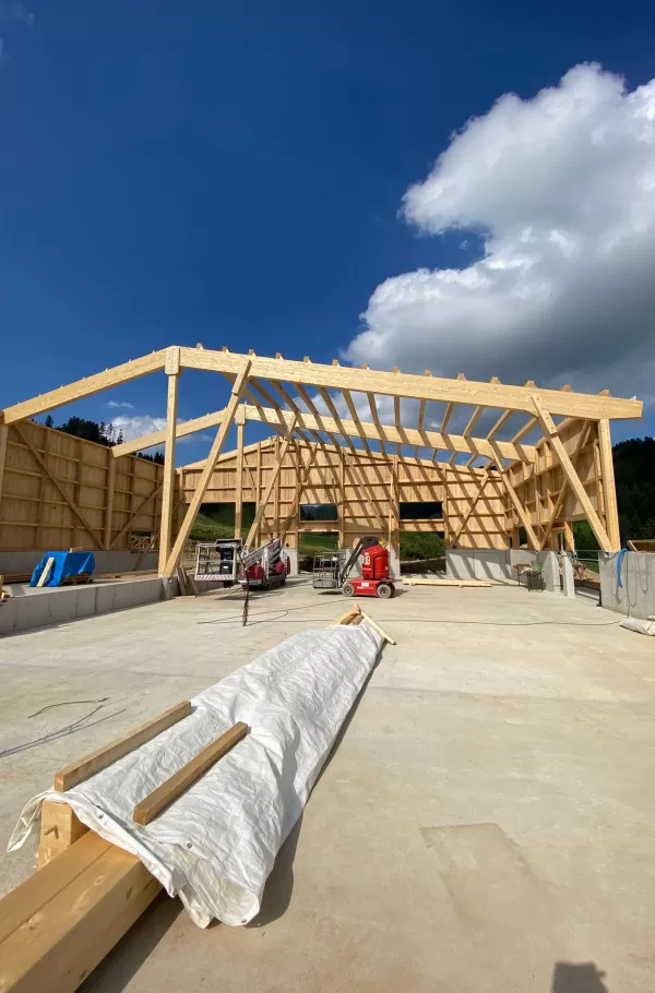 Rohbau einer großen Holzkonstruktion mit offenem Dachstuhl: Massive Holzträger und Streben formen die Grundstruktur des Gebäudes, während der freie Himmel mit weißen Wolken die Weite des Raumes unterstreicht. Auf dem Betonboden liegen Baumaterialien, während im Hintergrund Baugeräte bereitstehen. Das Bild zeigt die Dimension und Stabilität moderner Holzbauweise.