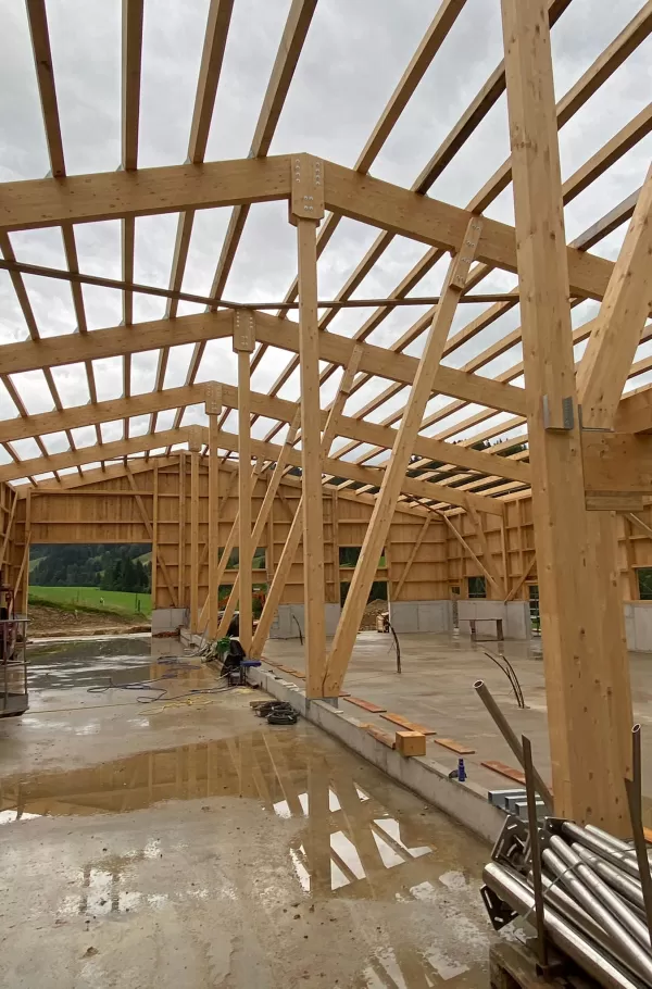 Innenansicht einer großen Holzkonstruktion im Rohbau: Stabile Holzträger und Streben formen das tragende Gerüst, das sich über die gesamte Gebäudefläche erstreckt. Auf dem Betonboden spiegeln sich die Strukturen im Regenwasser, während Werkzeuge und Baumaterialien den Baustellencharakter unterstreichen. Die Kombination aus Holz und Beton zeigt die Dimension und Präzision des modernen Holzbaus.