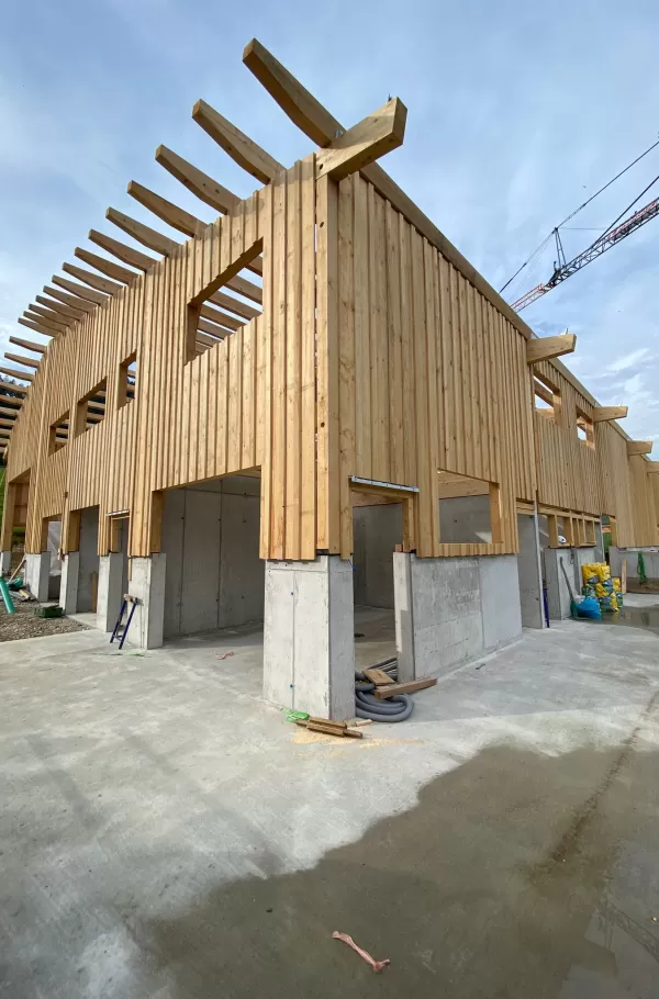 Detailansicht des Neubaus aus Holz und Beton: Die Fassade aus senkrechten Holzbrettern ist bereits montiert, während die massiven Betonstützen das Fundament und die Erdgeschossebene tragen. Sichtbare Dachbalken ragen über die Wände hinaus. Der Bau zeigt die Verbindung von moderner Stabilität mit traditioneller Handwerkskunst.