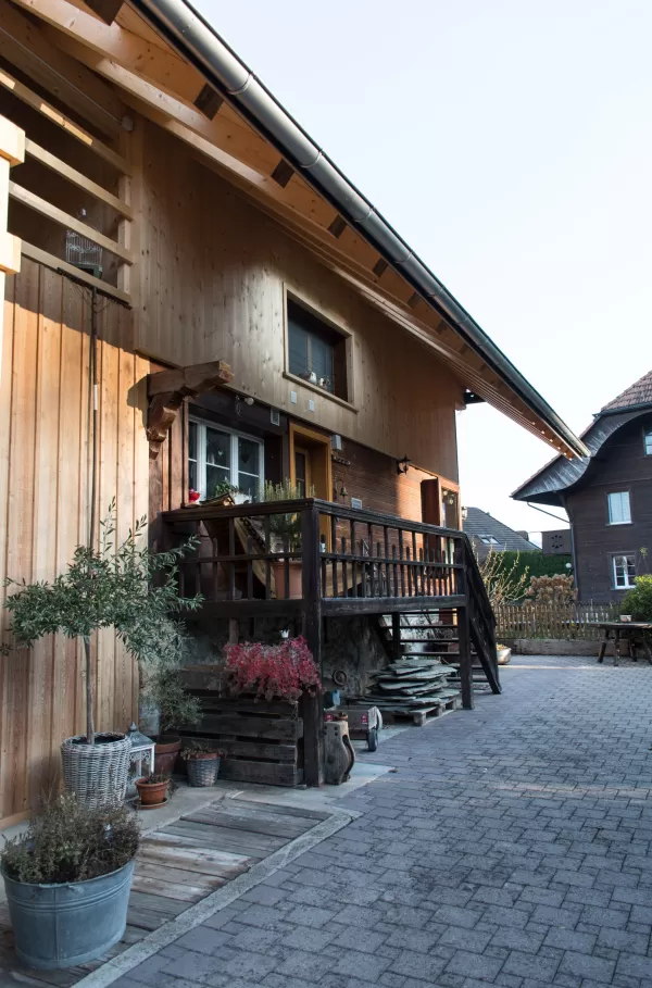 Holzhaus mit überdachtem Eingang und kleiner Veranda, umgeben von Pflanzgefäßen und gepflastertem Hof.