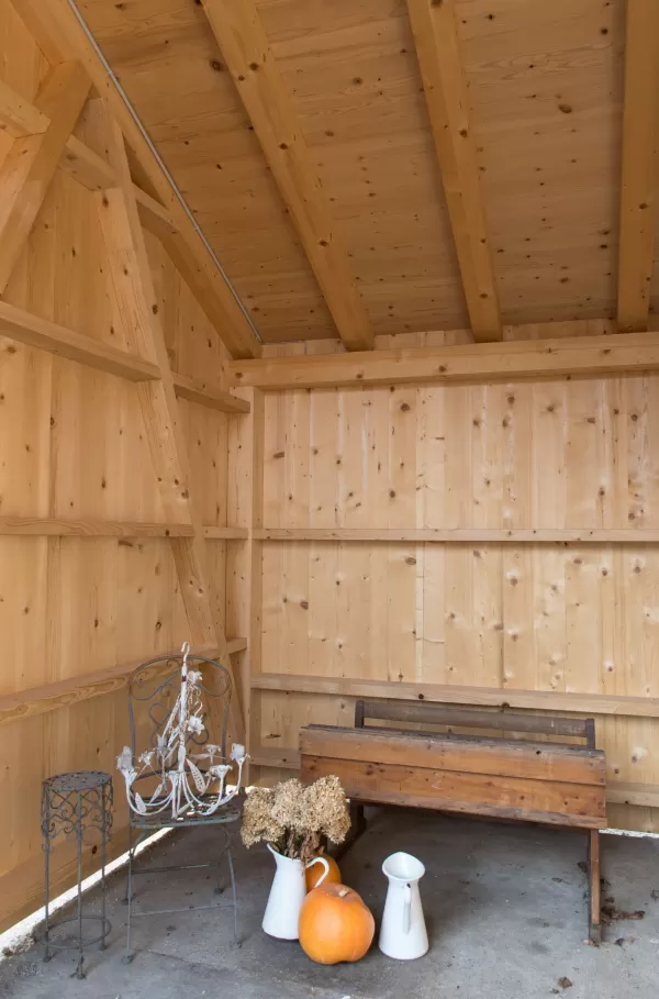 Überdachter Holzraum mit Bank, Metallstuhl, Kürbissen und getrockneten Blumen als herbstliche Dekoration.