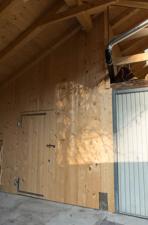 Innenansicht eines Holzbaus mit sichtbarer Balkenkonstruktion, Holzwand und kleiner Tür, teilweise im Sonnenlicht.