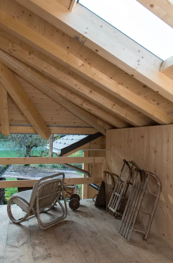Holzverkleideter Dachraum mit sichtbarer Balkenkonstruktion, Dachfenster, altem Schaukelstuhl und abgestellten Holzschlitten.