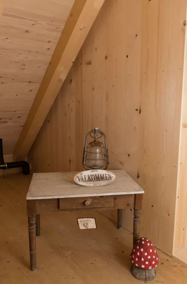 Kleiner Holztisch unter einer Dachschräge mit einer Lampe und einem Schild mit der Aufschrift ‚Välkommen‘, daneben eine rote Pilzdekoration.