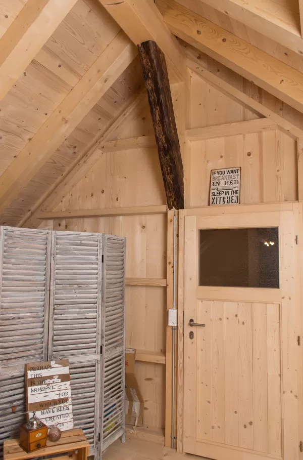 Innenansicht eines Dachraums mit Holzwänden, Tür mit Glasfenster, altem Holzbalken und grauem Paravent als Raumteiler.