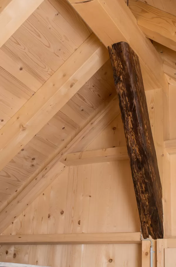 Detailansicht einer Dachkonstruktion aus hellem Holz mit einem dunklen, alten Balken als sichtbarem Stützelement.