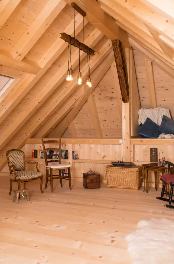 Gemütlicher Dachraum mit sichtbarer Holzbalkendecke, Holzboden und Einrichtung aus Stühlen, Bücherregal und warmem Licht.