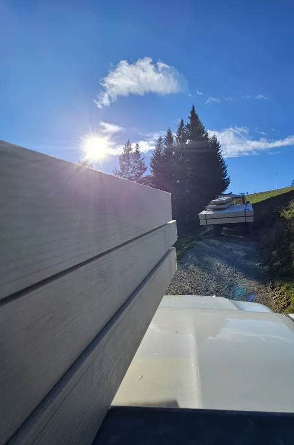 Montagearbeiten an einer Holzplattform vor einem traditionellen Gebäude auf einer Anhöhe. Ein Traktor mit Hebevorrichtung hebt einen langen Holzbalken an, während zwei Mitarbeitende der Zimmerei Hirschi AG den Balken ausrichten.