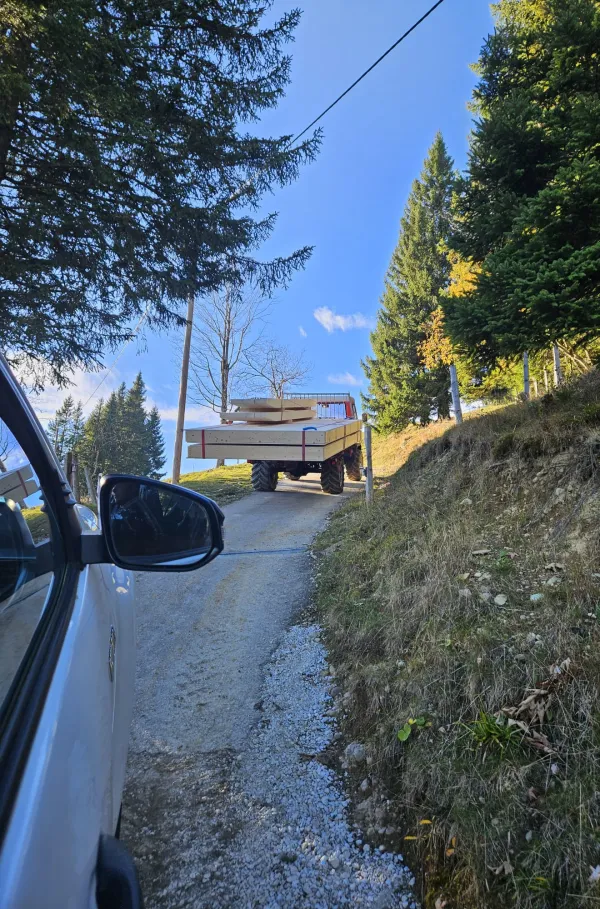Montagearbeiten an einer Holzplattform vor einem traditionellen Gebäude auf einer Anhöhe. Ein Traktor mit Hebevorrichtung hebt einen langen Holzbalken an, während zwei Mitarbeitende der Zimmerei Hirschi AG den Balken ausrichten.