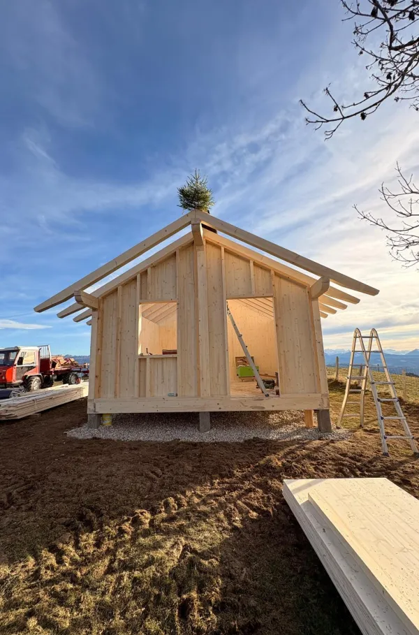 Montagearbeiten an einer Holzplattform vor einem traditionellen Gebäude auf einer Anhöhe. Ein Traktor mit Hebevorrichtung hebt einen langen Holzbalken an, während zwei Mitarbeitende der Zimmerei Hirschi AG den Balken ausrichten.