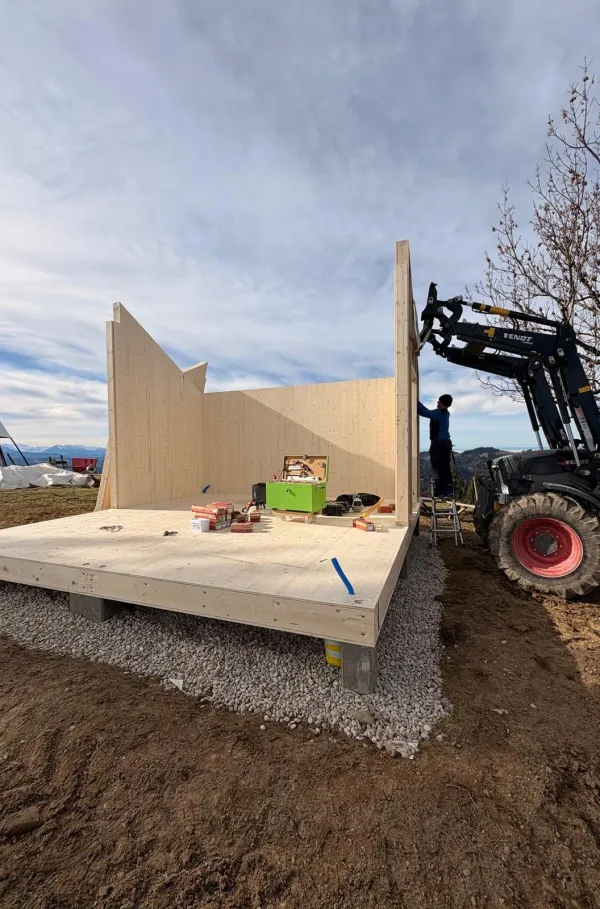 Montagearbeiten an einer Holzplattform vor einem traditionellen Gebäude auf einer Anhöhe. Ein Traktor mit Hebevorrichtung hebt einen langen Holzbalken an, während zwei Mitarbeitende der Zimmerei Hirschi AG den Balken ausrichten.