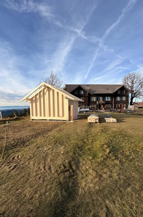 Montagearbeiten an einer Holzplattform vor einem traditionellen Gebäude auf einer Anhöhe. Ein Traktor mit Hebevorrichtung hebt einen langen Holzbalken an, während zwei Mitarbeitende der Zimmerei Hirschi AG den Balken ausrichten.