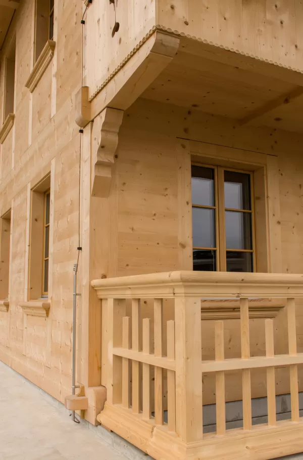 Detailaufnahme einer modernen Holzfassade mit Balkon und Fensterrahmen aus hellem Fichtenholz. Das Bild zeigt die präzise Zimmermannsarbeit mit sauber gearbeiteten Verbindungen und traditionellen Elementen.