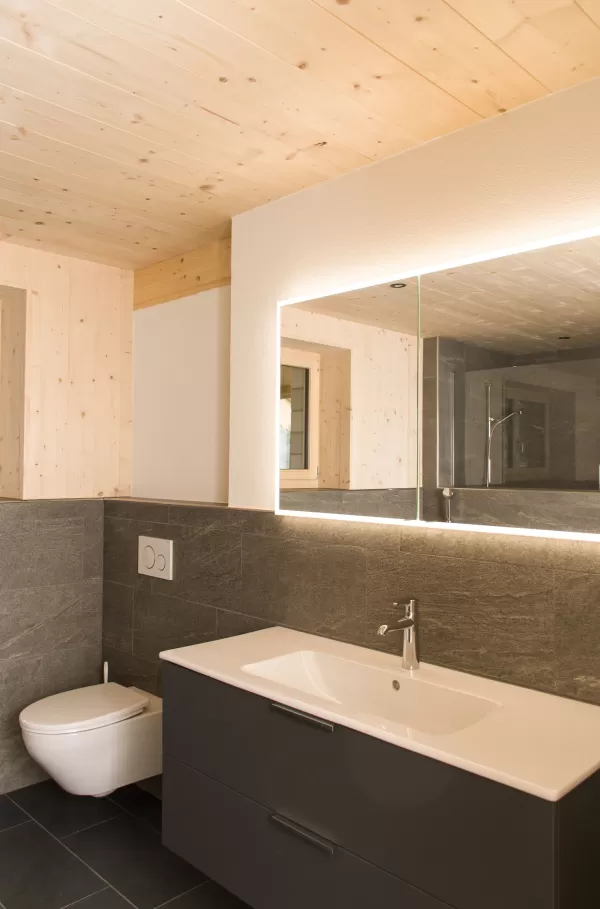Modernes Badezimmer mit Holzdecke und grauen Steinfliesen. Ein beleuchteter Spiegel, ein Waschtisch mit dunkler Front und eine wandhängende Toilette schaffen ein stilvolles, harmonisches Ambiente aus Holz und Stein.