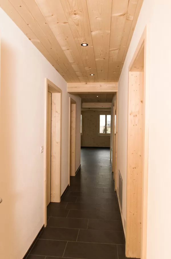 Heller Hausflur mit Holzdecke und Türrahmen aus Fichtenholz. Der dunkle Steinboden bildet einen eleganten Kontrast zur warmen Holzgestaltung und führt zu einem lichtdurchfluteten Raum im Hintergrund.
