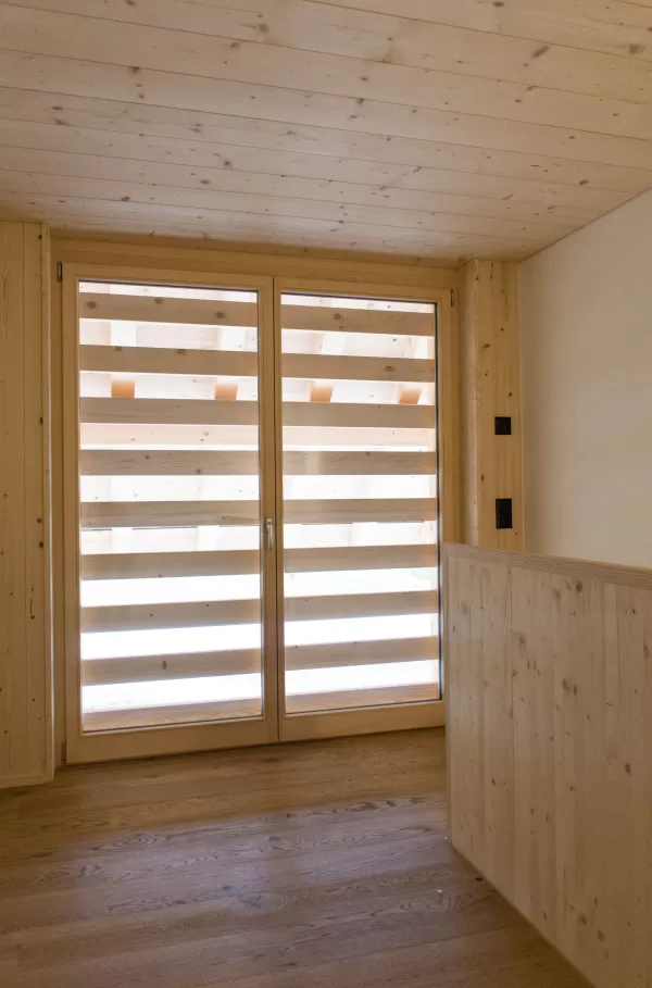 Raum mit Holzdecke und Boden aus Eichenholz, bodentiefem Fenster mit horizontalen Holzlamellen. Das natürliche Licht und die harmonische Holzgestaltung verleihen dem Raum eine warme, gemütliche Atmosphäre.