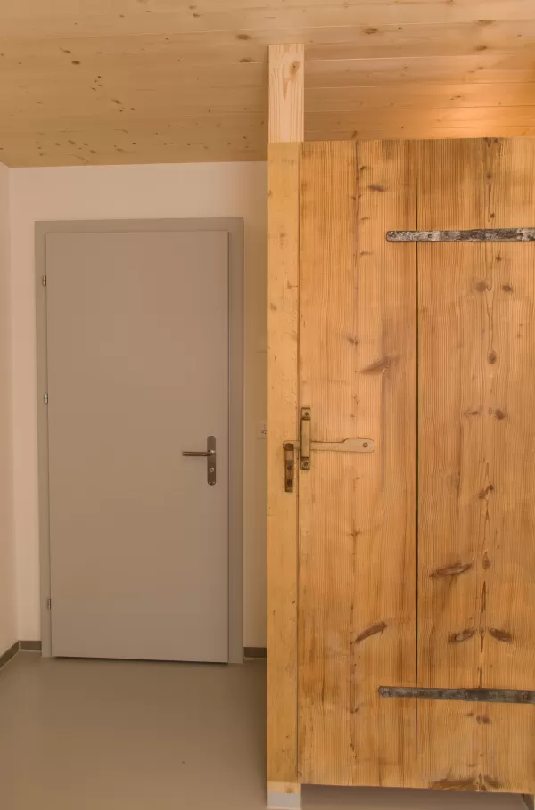 Innenraum mit modern-grauer Tür und daneben einer rustikalen Holztüre mit Eisenbeschlägen. Der Kontrast zwischen neuem und altem Holzhandwerk verleiht dem Raum einen besonderen Charme.