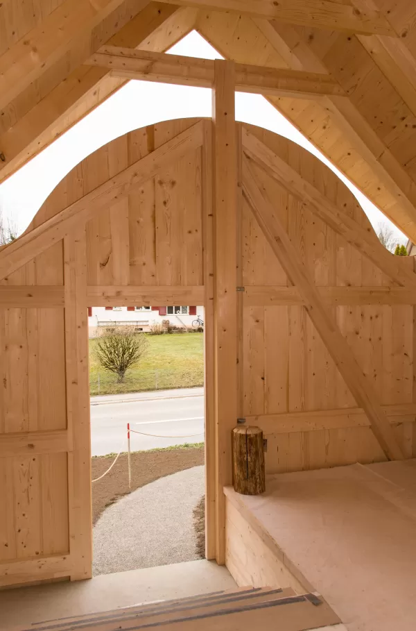 Innenansicht einer großen Holztoröffnung mit sichtbarer Dachkonstruktion. Durch die offene Tür fällt Tageslicht ein und gibt den Blick auf die angrenzende Straße und Wiese frei – ein Beispiel präziser Zimmermannsarbeit.
