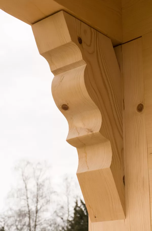 Detail einer kunstvoll gearbeiteten Holzkonsole unter einem Dachvorsprung. Die geschwungene Form und feine Verarbeitung betonen die traditionelle Handwerkskunst im modernen Holzbau.
