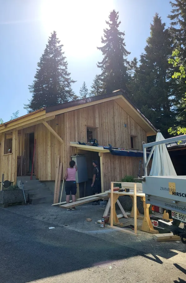 Holzhaus im Bau mit natürlicher Holzfassade inmitten einer Waldlandschaft. Das eingeschossige Gebäude steht auf einem gepflegten Grundstück mit Wiese und Zufahrtsweg. Die Fensteröffnungen sind bereits erstellt, während der Innenausbau noch im Gange ist. Im Hintergrund befinden sich weitere Holzbauten sowie hohe Nadelbäume, die das Gebäude umgeben. Die Aufnahme zeigt die harmonische Einbindung des Holzbaus in die natürliche Umgebung.
