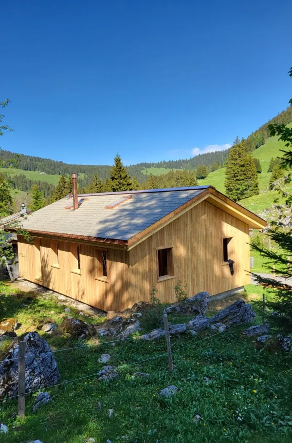 Holzhaus im Bau mit natürlicher Holzfassade inmitten einer Waldlandschaft. Das eingeschossige Gebäude steht auf einem gepflegten Grundstück mit Wiese und Zufahrtsweg. Die Fensteröffnungen sind bereits erstellt, während der Innenausbau noch im Gange ist. Im Hintergrund befinden sich weitere Holzbauten sowie hohe Nadelbäume, die das Gebäude umgeben. Die Aufnahme zeigt die harmonische Einbindung des Holzbaus in die natürliche Umgebung.